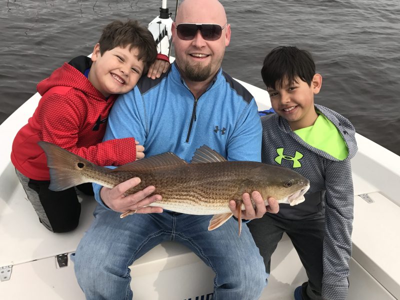 Man and two children on a boat holding a fish and smiling at the camera.