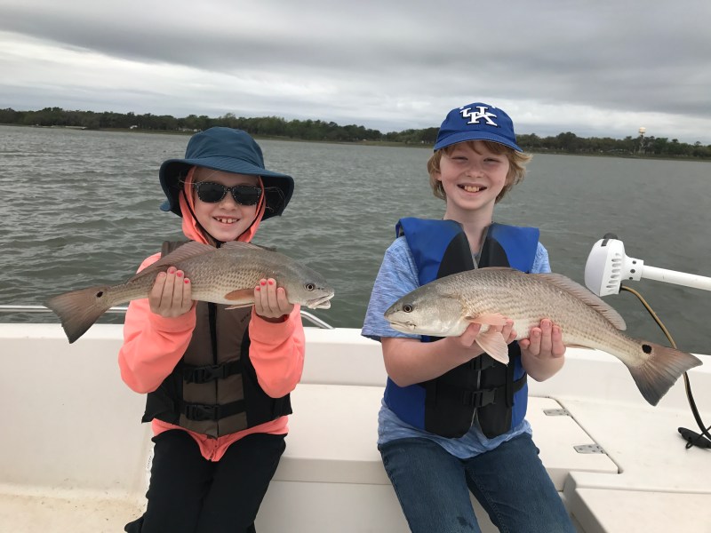 Two children on a boat holding fish, wearing life vests and smiling.
