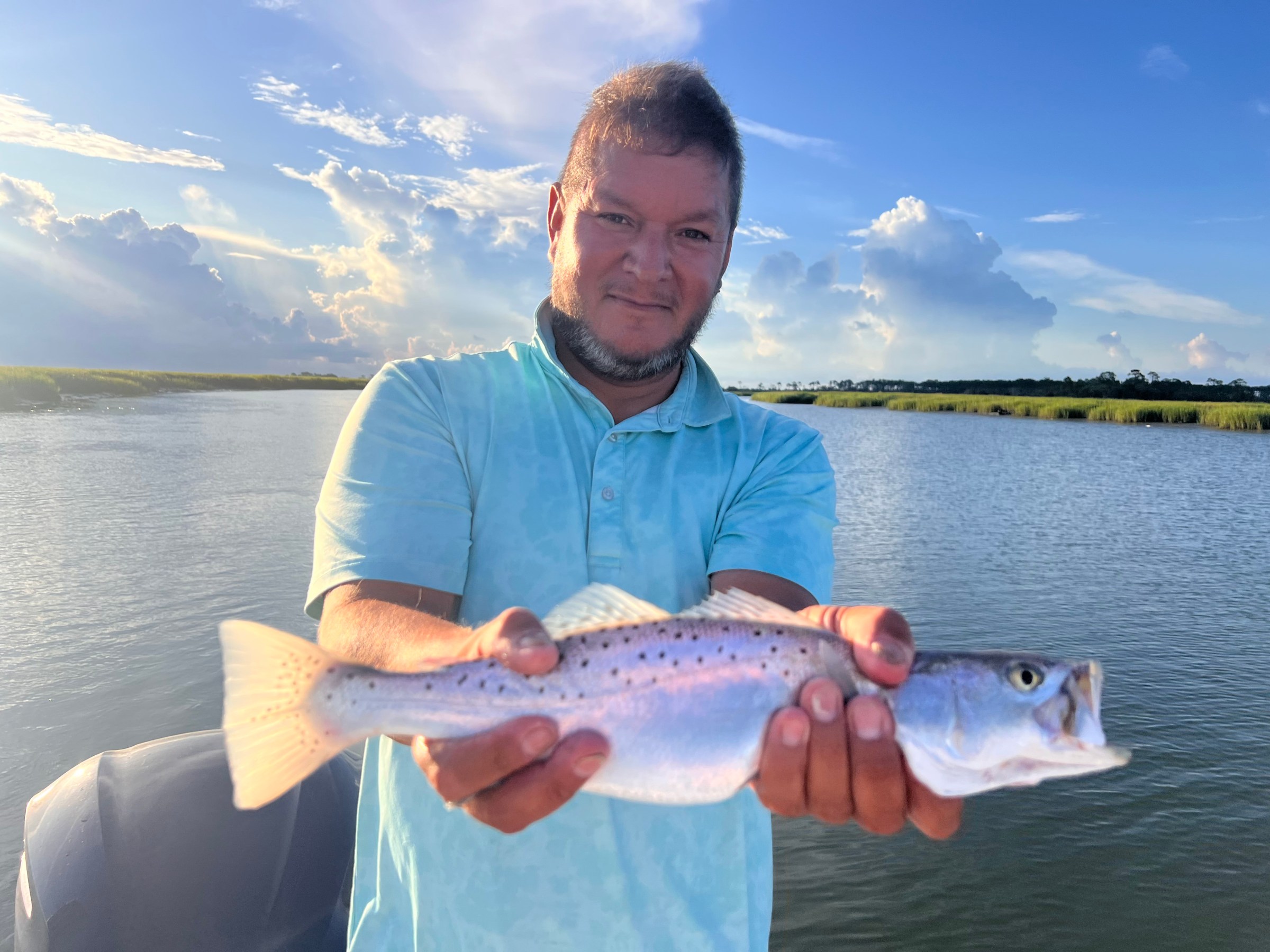 Person holding a fish on a boat with a scenic river and cloudy sky background.