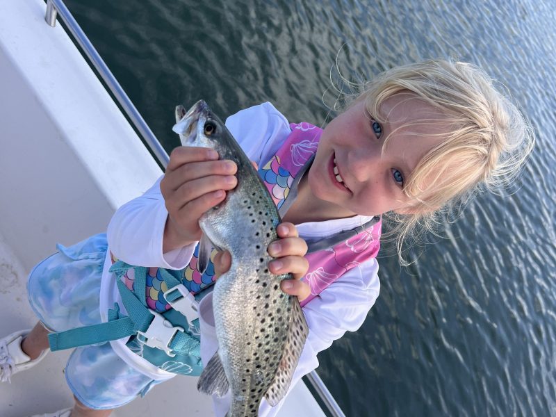 Child on a boat holding a fish, smiling with water in the background.