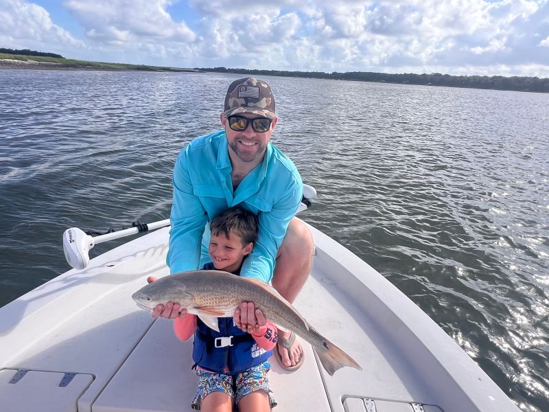 Man and child on boat holding a fish with water in the background.