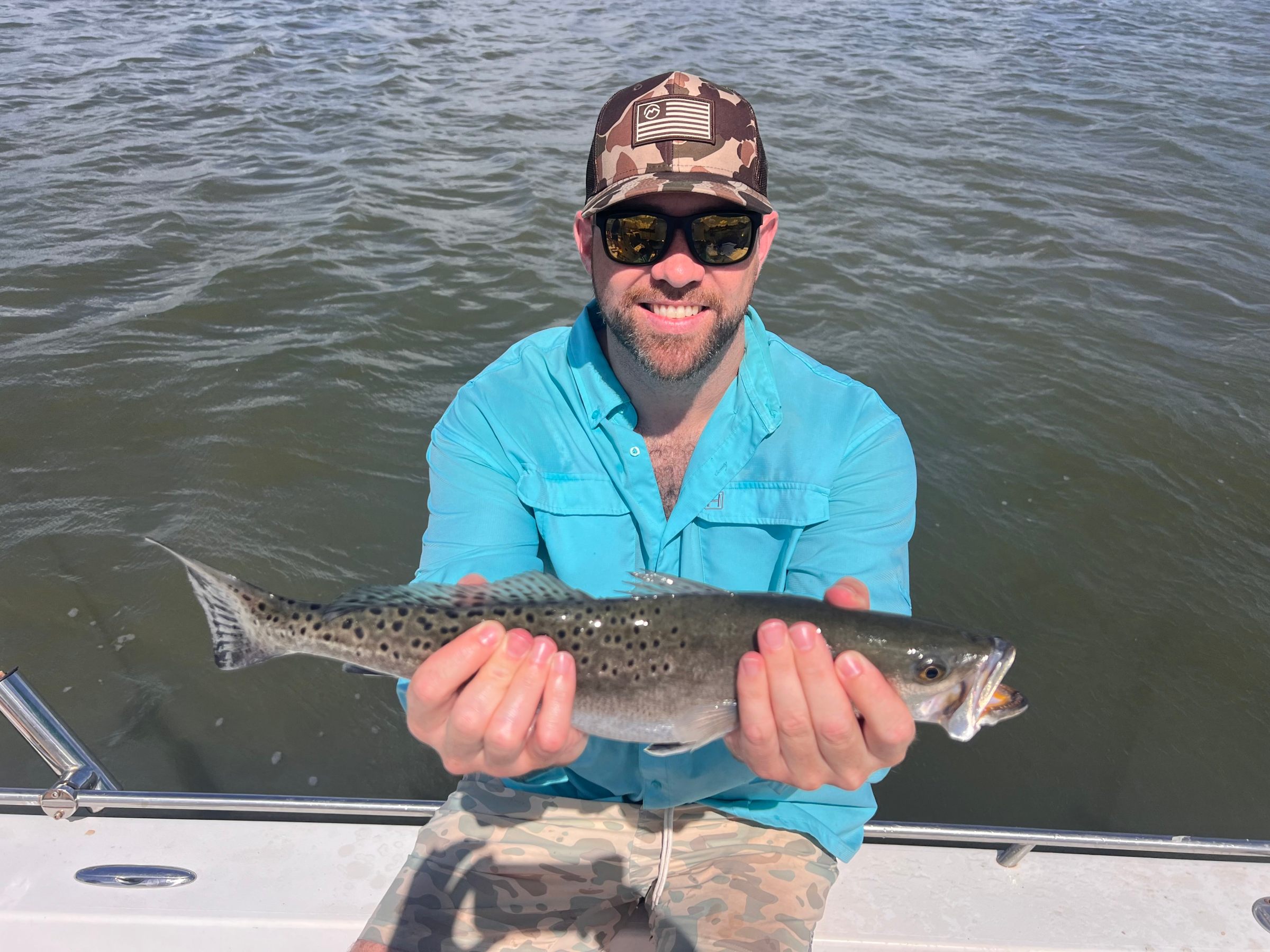 Man in blue shirt and camo hat holds a fish on a boat with water in the background.