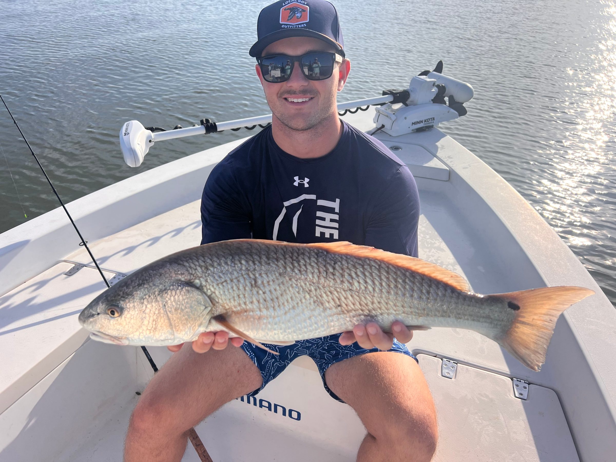 Person in a boat holding a large fish, smiling, wearing sunglasses and a hat.