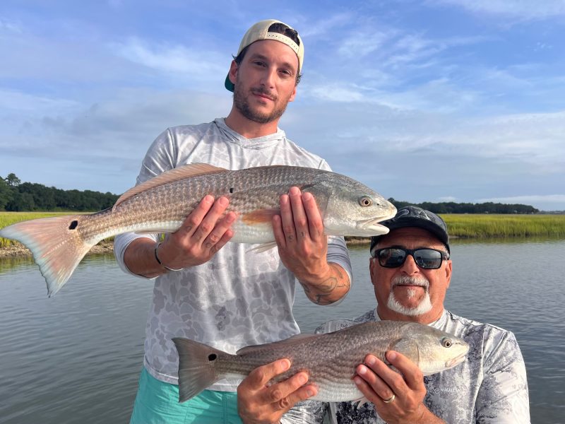 Two men hold fish on a boat near marshy water.