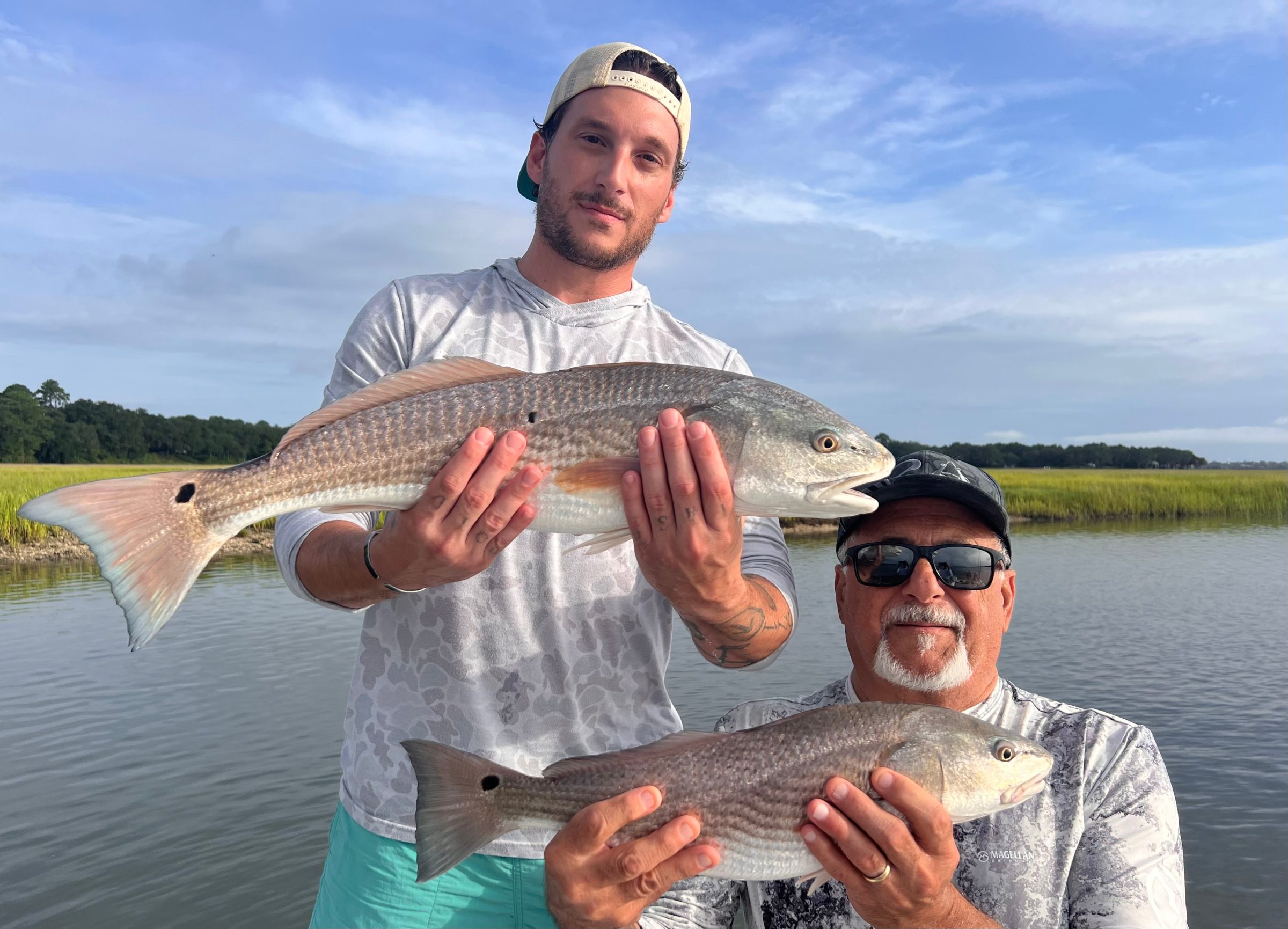 Two men hold fish on a boat near marshy water.