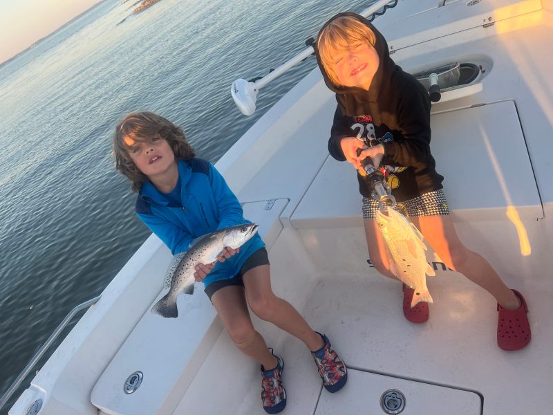 Two children on a boat holding fish, with water in the background.