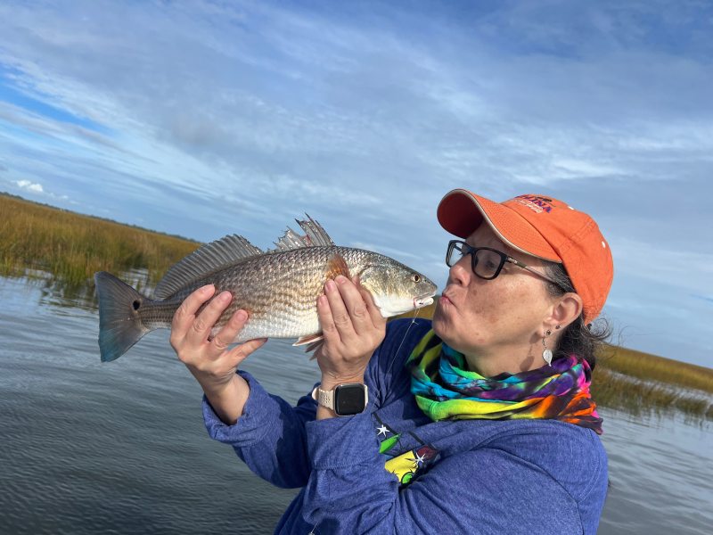 Person kissing a fish near water with reeds in the background.