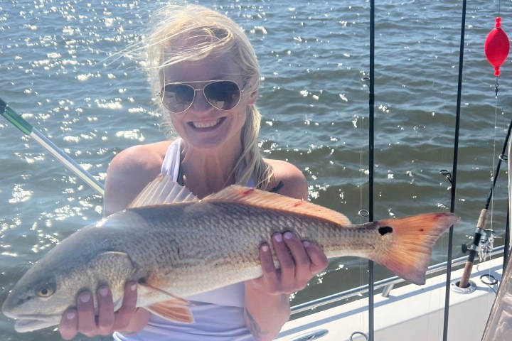 Person in white tank top holds fish on a boat, with water and fishing gear in background.