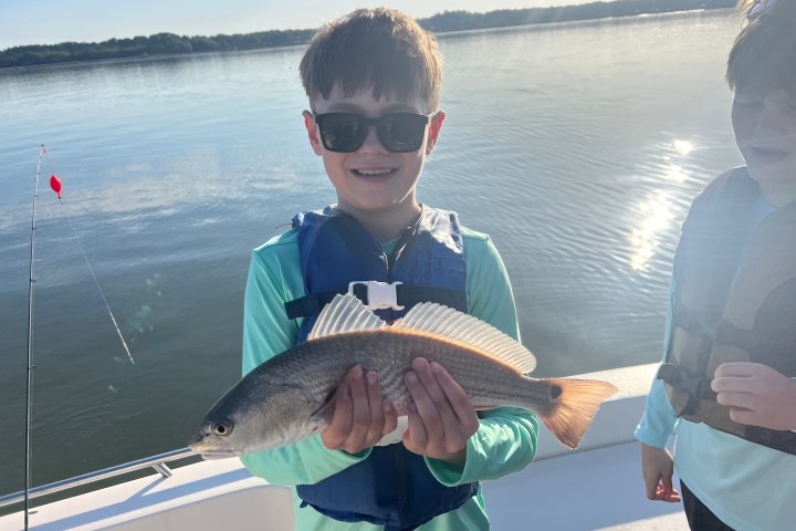 Boy in sunglasses holding a fish on a boat, with a calm lake in the background.