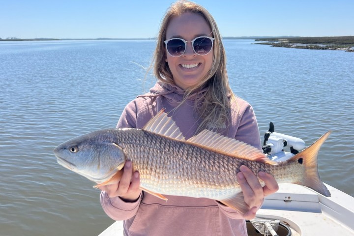 Person in sunglasses holding a fish on a boat with a calm water background.